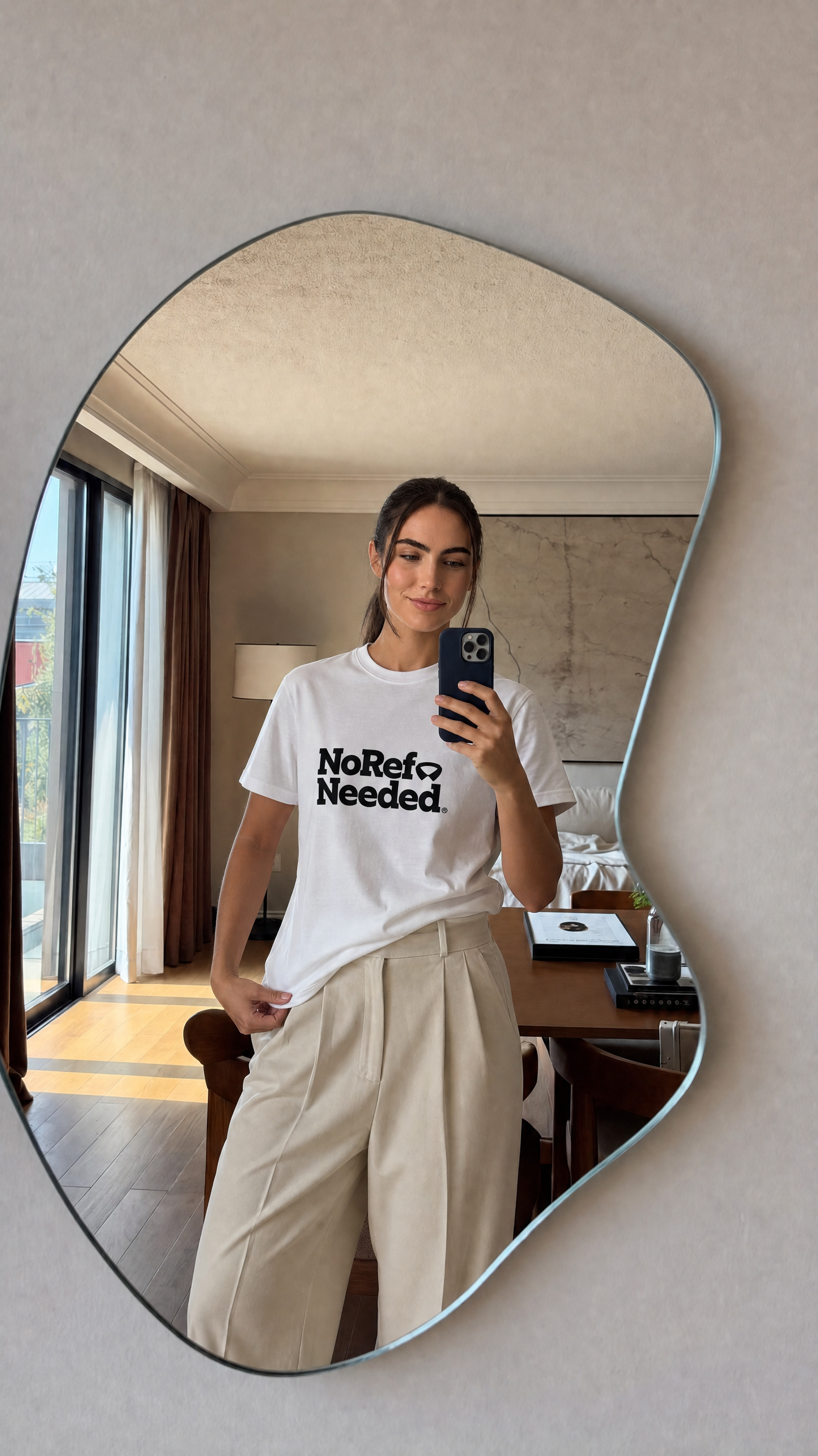 Person taking a mirror selfie wearing a white t-shirt with text in a modern interior setting.